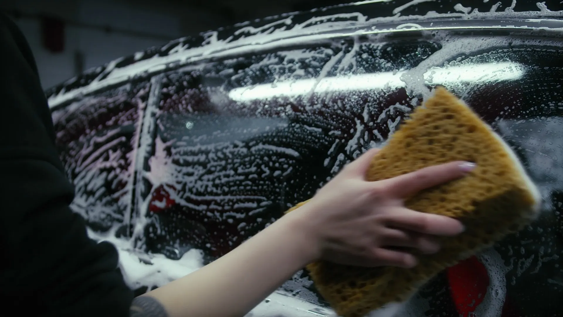 Mão com esponja espalhando espuma sobre o vidro de um carro escuro